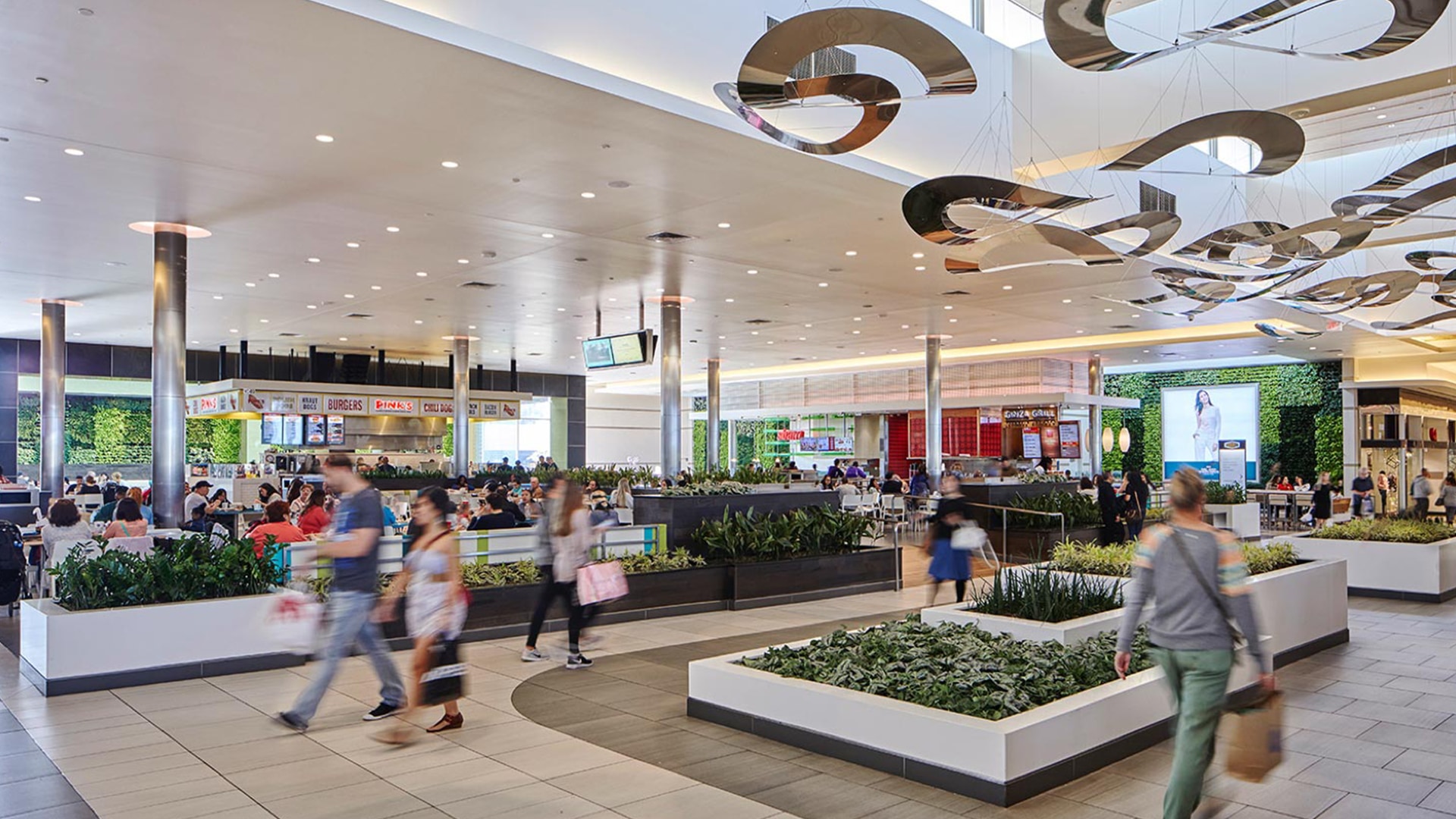 Inside a Simon Malls Food court, in the background groups sit a tables surrounded by numerous food storefronts and pedestrians walk by the decorated center aisles.